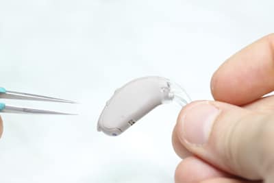 A closeup of a person holding a hearing aid in one hand and a set of tweezers in the other