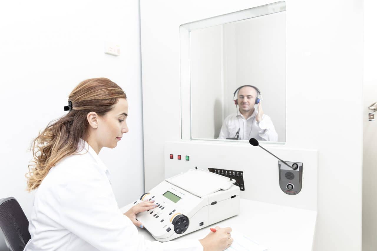 female audiologist testing a patient's hearing while male patient sits in booth