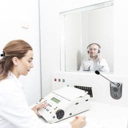 female audiologist testing a patient's hearing while male patient sits in booth